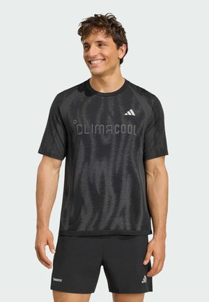 Homme souriant portant un t-shirt noir Adidas Climacool à manches courtes et un short assorti sur un fond clair uni.