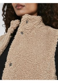 Béžová fleecová vesta s límcem, vyrobená z měkké, strukturované tkaniny se šedými cvoky. Nositelná přes černý top, vytváří vrstvený vzhled.