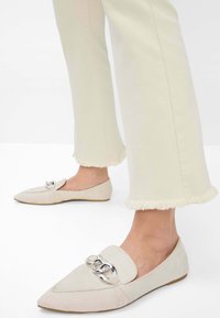 Beige suede loafers with a pointed toe, featuring a silver chain accent and a flat sole. Paired with light-colored, frayed hem trousers.