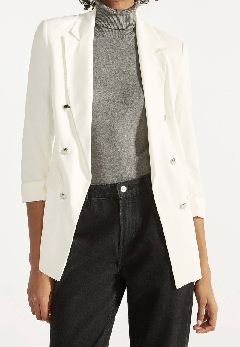 Blazer blanc à double boutonnage avec boutons en argent, coupe ajustée et manches retroussées, associé à un col roulé gris et un pantalon noir.