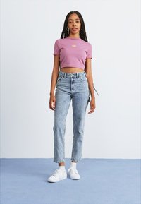 Ung kvinne med lange fletter iført en rosa cropped t-skjorte, lyseblå høytlivsjeans, hvite sokker og hvite joggesko står på blått gulv.