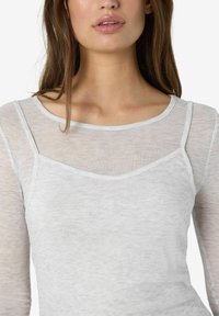 Sieviete ar gariem brūniem matiem, tērpusies baltā ribotā daudzslāņu blūzē ar caurspīdīgām garām piedurknēm un noapaļu kakla izgriezumu.