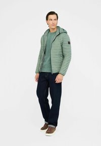 Lysgrøn pufferjakke med hætte, lynlås foran og sidelommer, stylet med en struktureret grøn sweater og mørke jeans. Brune sneakers med snørebånd.