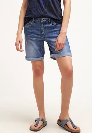Personne portant un short en denim bleu retroussé, un haut bleu marine et des sandales en liège bleu foncé, debout devant un fond uni.