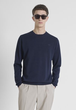 Maglione lavorato a maglia blu navy con scollatura rotonda e maniche lunghe. Presenta un logo discreto sul petto, abbinato a pantaloni beige.