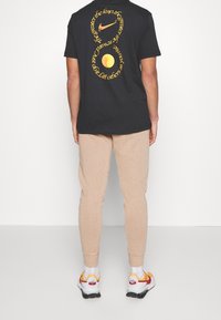 Svart bomullströja med gul text och logotyp på ryggen, som matchas med beige sweatpants och flerfärgade sneakers.