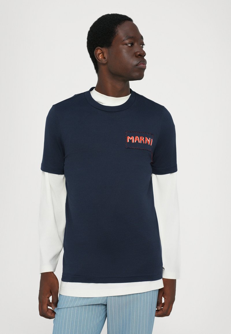 Camiseta de manga corta de color azul marino con una capa blanca de manga larga debajo, que presenta un bolsillo con logo bordado rojo en el pecho. Material suave de algodón.