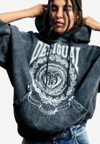 Czarna, oversize bluza z kapturem ze ściągaczem, z białym emblematem i ozdobnymi grafikami na przodzie. Miękka, teksturowana bawełniana tkanina.