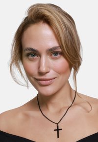 Collana con pendente a forma di croce nera su una catena sottile, indossata con un top off-shoulder. Trucco naturale e capelli sciolti incorniciano il viso.