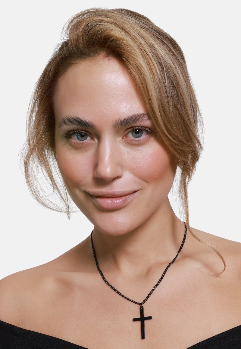 Collana con pendente a forma di croce nera su una catena sottile, indossata con un top off-shoulder. Trucco naturale e capelli sciolti incorniciano il viso.