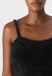 Ingrandimento di una persona che indossa un top senza maniche nero e peloso con spalline sottili e textured, su uno sfondo bianco semplice.