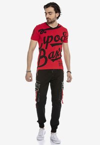 Rotes Baumwoll-T-Shirt mit großen schwarzen Schriftzügen; kombiniert mit schwarzen Cargo-Hosen mit Reißverschlusstaschen und roten Akzenten. Weiße Sneaker.