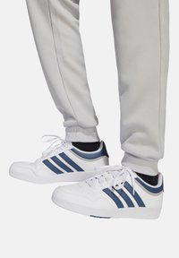 Weiße Sneakers mit blauen Akzenten, ausgestattet mit drei Streifen, glattem Lederobermaterial, runder Zehenpartie und Schnürdesign. Getragen mit grauen, schmal zulaufenden Hosen.