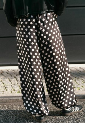 Personne portant un pantalon noir ample à gros pois blancs et des baskets noires, debout sur une surface pavée en plein air.