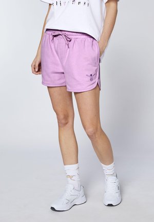 Person trägt lila Shorts mit Kordelzug und einer kleinen eingestickten Figur, weißes bedrucktes Shirt, weiße gemusterte Socken und weiße Sneakers.