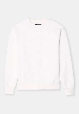 Henkilö, jolla on yllään valkoinen pitkähihaisen collegepaita, harmaat urheilushortsit ja valkoiset lenkkitossut, seisomassa harmaalla lattialla.