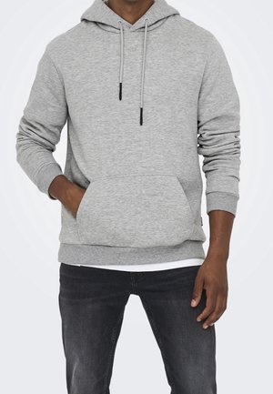 Mand iført lys grå hættetrøje med frontlomme og sorte jeans, står med den ene hånd i lommen mod en ensfarvet baggrund.