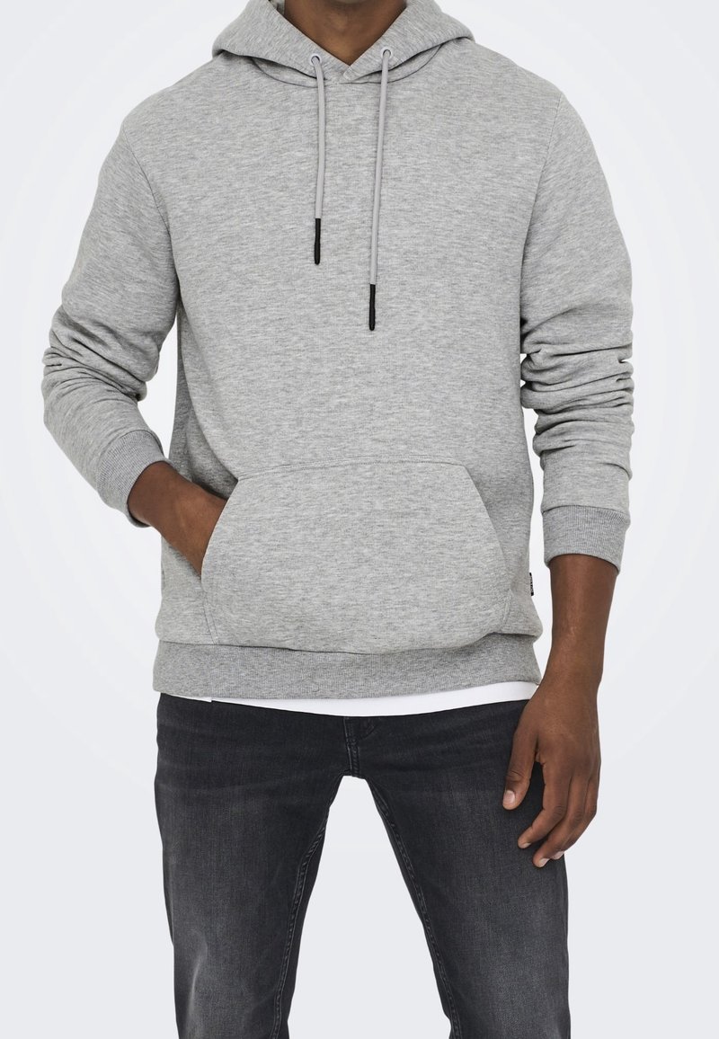 Homme portant un sweat à capuche gris clair avec poche avant et jean noir, debout avec une main dans la poche devant un fond uni.