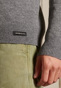 Pull gris côtelé avec une étiquette noire portant l'inscription "SUPERDRY." Main visible reposant sur un pantalon vert à motif à carreaux et texture élégante.