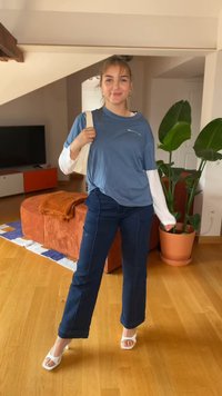 Sinine T-särk valge pika varrukaga pluusi peal, koos tumesiniste pükste ja valgete sandaalidega. Siseruum, kus on taimi ja mööblit.