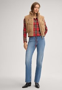 Gilet sherpa tan su una camicia a quadri rossa, abbinato a jeans dritti blu e stivaletti neri alla caviglia. Presenta una tasca con zip sul gilet.