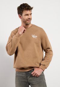 Barna pulóver puha anyagból, kerek nyakkivágással és hosszú ujjakkal. "Friss Kávé" felirat fehér színben a bal mellen. Lazán illeszkedő fazon.