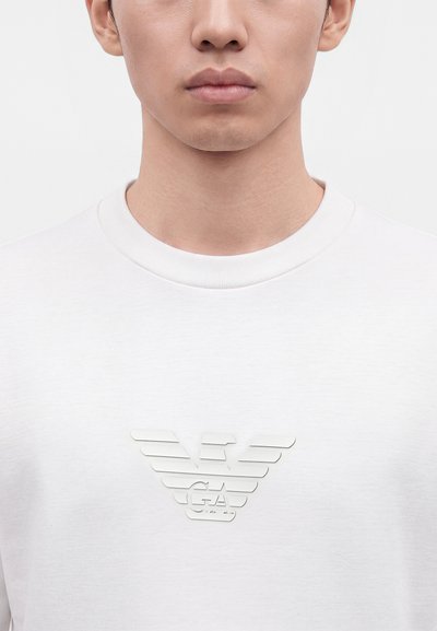 Gros plan d'une personne portant une chemise blanche avec un logo aigle stylisé en relief blanc et les lettres G A sur la poitrine.