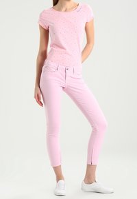 Žena oblečená v světle růžových slim džínách s zipy na kotnících, odpovídajícím krátkým růžovým tričku s puntíky a bílých teniskách.