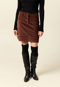 Ruskea brokadihame, jossa on etupainike, taskut ja kapea malli. Yhdistetty mustiin polven yli ulottuviin saappaisiin ja tekstuurisiin mustiin sukkiin.