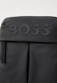 Sac à dos en tissu noir avec une surface texturée, logo "BOSS" en relief à l'avant, et un design épuré avec des détails de pliage.