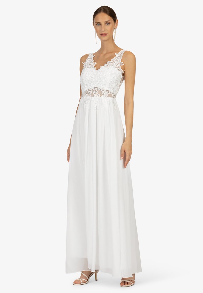 Weißes Kleid mit einem taillierten Spitzenoberteil mit floralen Details und einem fließenden Rock. Das Kleid hat einen V-Ausschnitt und durchsichtige Akzente. Sandalen sichtbar.