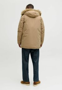 Giacca piumino beige con cappuccio foderato in pelliccia, caratterizzata da un taglio dritto e maniche lunghe, abbinata a pantaloni di denim scuro e scarpe marroni.