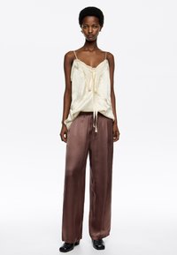 Camiseta de seda crema con encaje y detalle de lazo en la parte delantera, combinada con pantalones de satén marrón de tiro alto y pierna ancha, y zapatos negros.