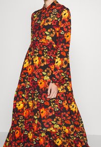 Šaty s dlouhým rukávem v černé látce, které mají živý květinový vzor v červené, oranžové a žluté barvě, s zapínáním na knoflíky u límce a vrstvenou sukní.