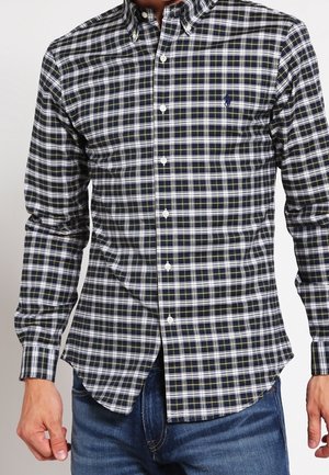 Homme portant une chemise à carreaux noire, blanche et jaune à manches longues et un jean bleu sur un fond uni.