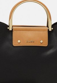 Melnas ādas rokassoma ar gaiši brūnu vāciņu, uz kura redzams zelta logo. Zelta metāla detaļas un noapaļota roktura dizains padara somu elegantāku.