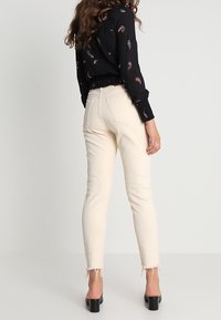Cream-colored skinny jeans with frayed hems, made from denim. Worn with a black patterned blouse and high-heeled shoes, viewed from the back.