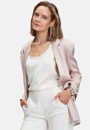 Frau trägt ein hellrosa Blazer über einer weißen Bluse und Hose, die Hände in den Taschen, dazu silberne Halskette, Ohrringe und Uhr als Accessoires.