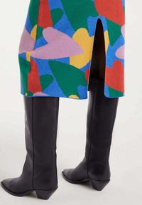 Botas de cuero negras hasta la rodilla, usadas con una falda de patrón abstracto colorido que presenta una abertura en la parte trasera.