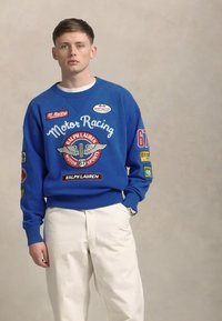 Blauwe sweatshirt met een "Motor Racing" grafiek en verschillende patches, gecombineerd met witte broek. Zachte katoen, ronde hals ontwerp.