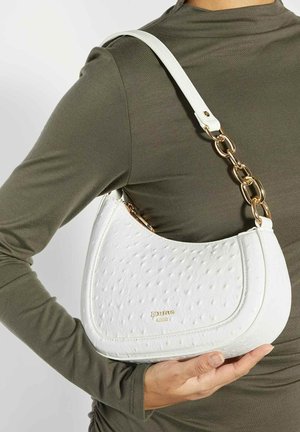 Witte gestructureerde handtas met gouden kettingband, vastgehouden door een persoon die een olijfgroene top met lange mouwen draagt, tegen een effen achtergrond.