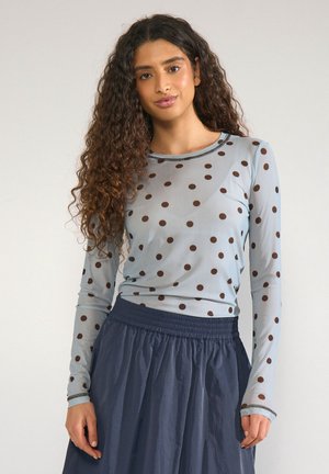 Kvinde med langt krøllet hår iført en lys blå bluse med brun polkaprikket og en mørkeblå samlet nederdel.