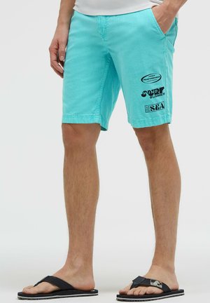 Man wearing turquoise shorts with black summer-themed text on right leg and black flip-flops, standing with hands in pockets.