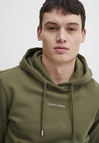 Olívazöld kapucnis pulcsi puha anyagból, elől kenguru zsebbel, húzózsinórral és "CASUAL FRIDAY" felirattal.