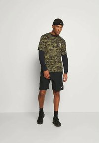 Camo-mønstret sportstrøje i grøn og sort, lagd over en langærmet sort skjorte, parret med sorte shorts og sorte sneakers.