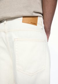 White denim jeans with a textured finish, featuring a tan leather patch labeled "PULL&BEAR" and standard back pocket design.