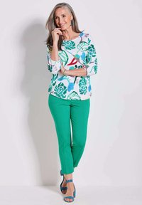 Bloemenprint top met groene, blauwe en rode accenten, gecombineerd met groene slim-fit broek. Model draagt blauwe hoge hakken. Eenvoudige achtergrond.
