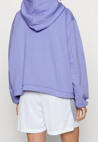 Violetti huppari, jossa on rento istuvuus, pehmeä materiaali ja huppu. Yhdistetty valkoisiin shortseihin, joissa on harmaa aksentti sivussa.