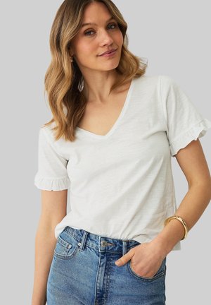 Kvinde med bølget blondt hår iført en hvid V-hals T-shirt med flæseærmer og blå jeans, med den ene hånd i lommen og en guldbøjle om håndleddet.