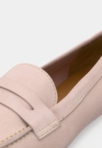 Mocassins en suède rose clair avec un bout arrondi, des coutures décoratives et un accent à onglet sur le cou-de-pied. L'intérieur présente une doublure jaune contrastante.